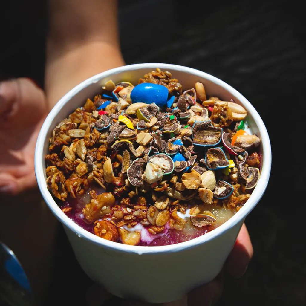 A thick deep purple açaí smoothie bowl in a wide white ceramic bowl, topped with neat rows of sliced kiwi, fresh strawberries, blueberries, granola, and a drizzle of honey. Bright clean natural daylight. Top-down shot on a white marble surface with a vibrant, colorful food photography style.