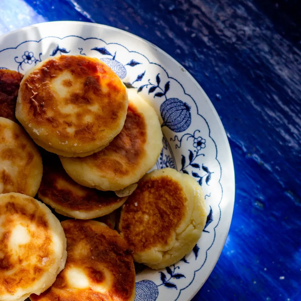 A stack of four golden cottage cheese pancakes on a white ceramic plate, topped with a pat of butter, a drizzle of warm maple syrup, and fresh blueberries scattered on top. The pancakes have a light, fluffy texture with visible golden edges. Warm morning kitchen light. 45-degree angle.