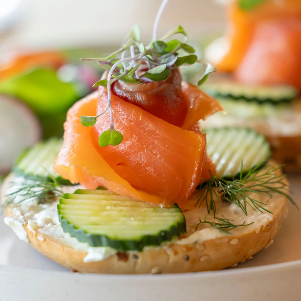 An open-face whole grain bagel on a white ceramic plate, spread with cream cheese and layered with silky smoked salmon, thin red onion rings, capers, and fresh dill. A lemon wedge sits beside it. Bright natural daylight from the side. 45-degree angle on a pale marble surface.