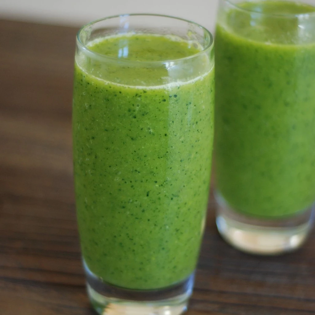 A vibrant bright green smoothie in a clear glass on a white marble counter, topped with a thin cucumber slice on the rim and a sprinkle of hemp seeds. Fresh spinach leaves and a cut lemon sit beside it. Clean bright morning daylight. Top-down food photography angle with a fresh, clean aesthetic.