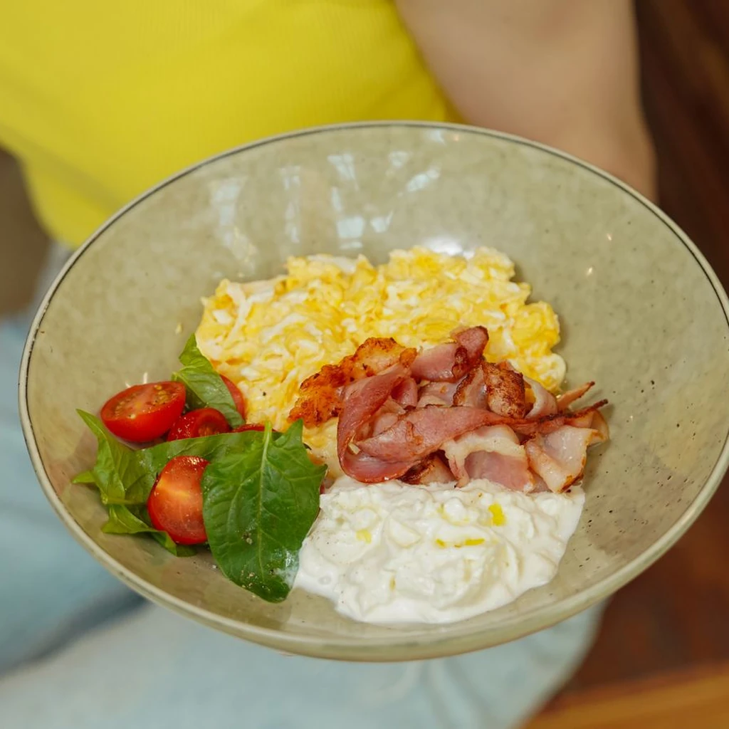 A rustic white bowl of fluffy scrambled eggs with ground turkey, diced tomatoes, sautéed spinach, and feta crumbles, garnished with fresh parsley. The egg texture is soft and creamy with visible vegetable pieces. Warm overhead kitchen light. 45-degree angle on a dark wood surface.