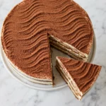 Overhead view of whole tiramisu cake on white cake stand with cocoa-dusted surface and one slice pulled away on white marble